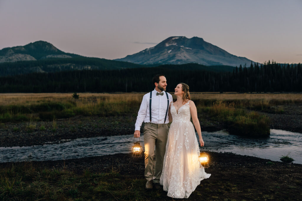 A bride is a blush lace gown smiles at her groom in a white shirt with suspenders and a bowtie carrying lanterns in the pre-dawn hour at their Sparks Lake Elopement in Bend, Oregon. 