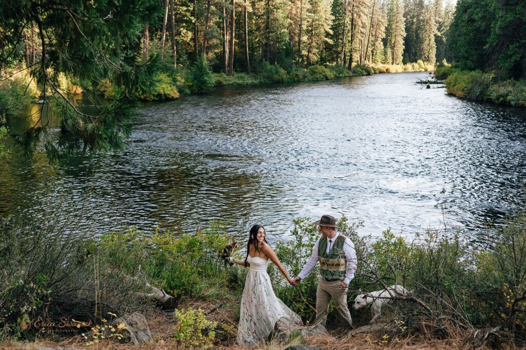 Bride and groom holding hands while walking by the Metolius River at their campground wedding, surrounded by lush forest.