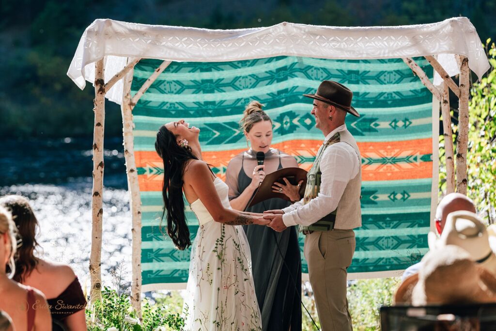 Bride laughing joyfully while holding hands with groom during their Metolius River wedding ceremony under a colorful blanket backdrop.