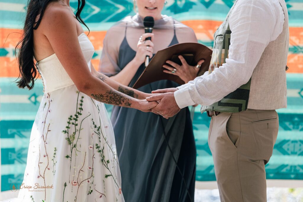 Bride and groom holding hands during their Metolius River wedding ceremony under a colorful blanket backdrop.