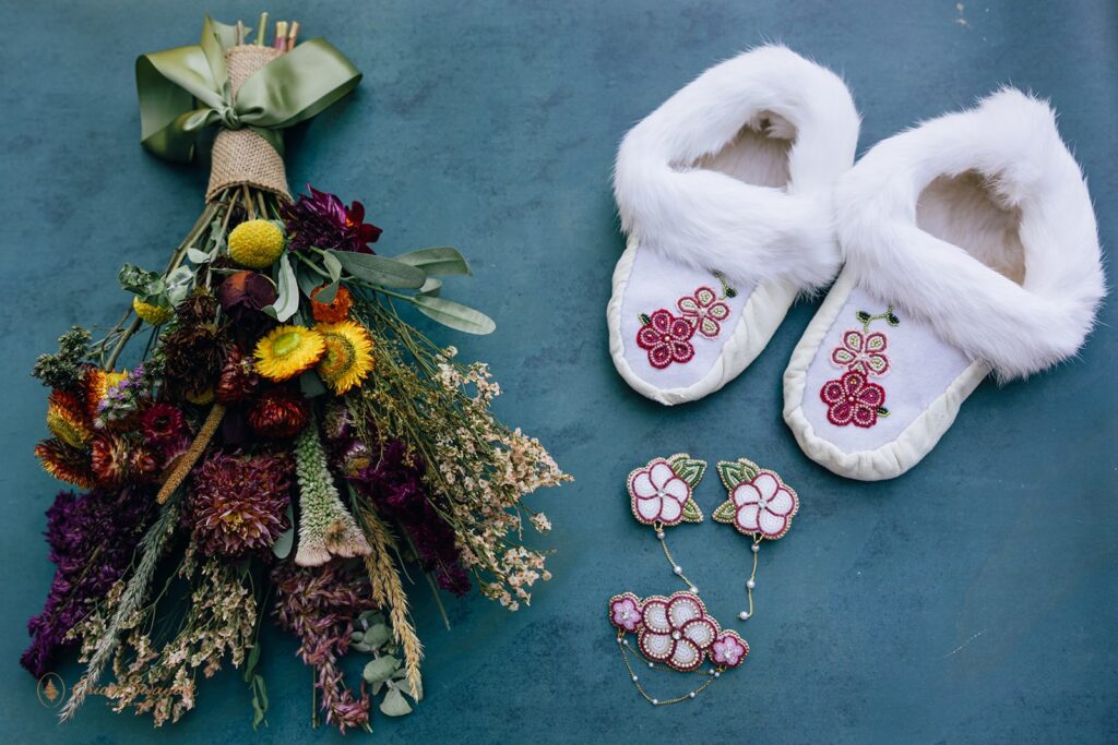Flat lay of bridal details with colorful bouquet, beaded floral jewelry, and white fur-trimmed moccasins with beadwork.