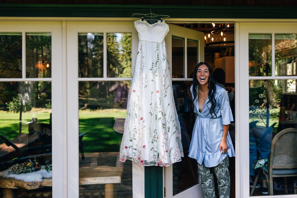 Bride laughing in a blue silk robe while standing beside her floral embroidered wedding dress hanging on a cabin door.