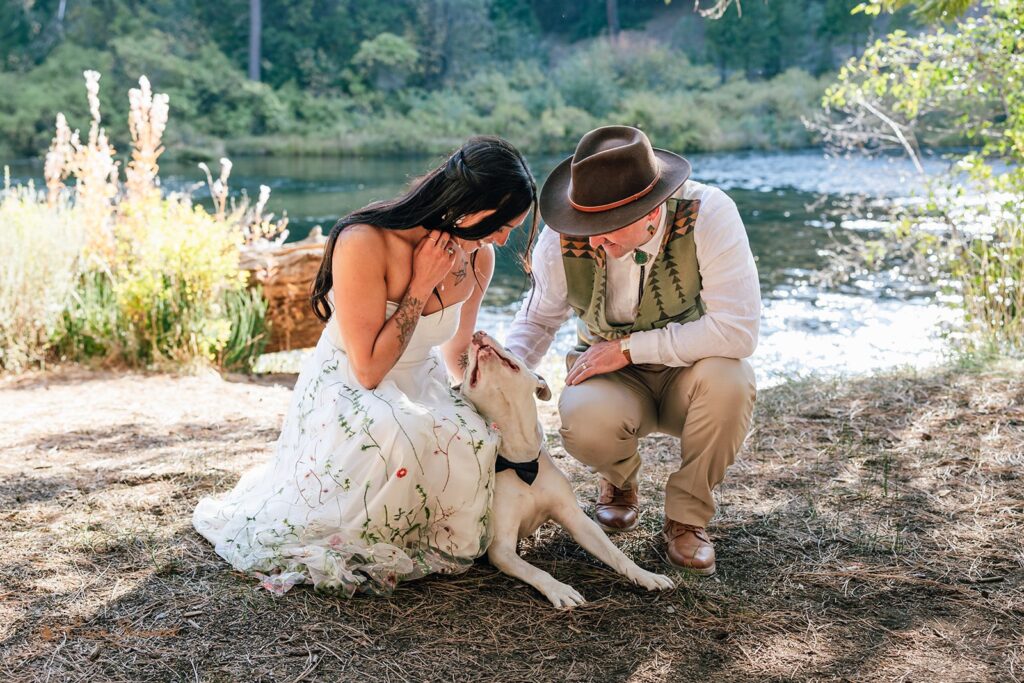 Bride and groom kneeling beside their dog by the Metolius River, the bride laughing as the pup gives her a kiss.