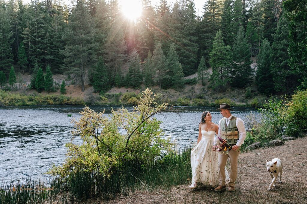 Bride and groom holding hands while walking by the Metolius River at their campground wedding with their dog