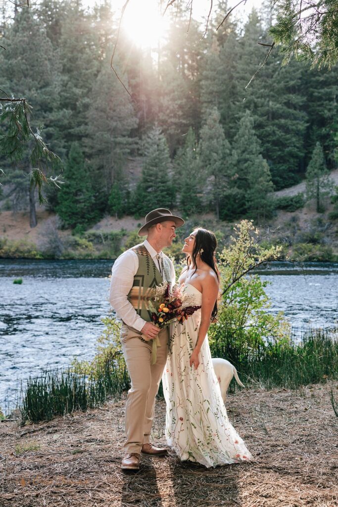 Bride and groom looking at each other while walking by the Metolius River at their campground wedding, surrounded by lush forest.