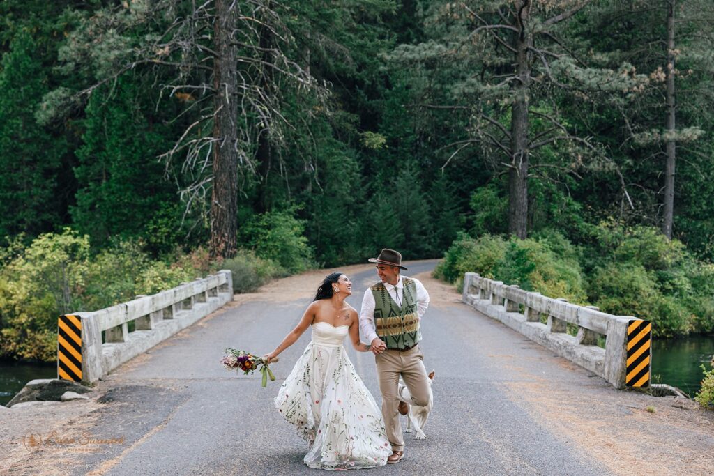 Newlyweds running hand in hand across a rustic bridge in the forest during their Metolius River wedding celebration.