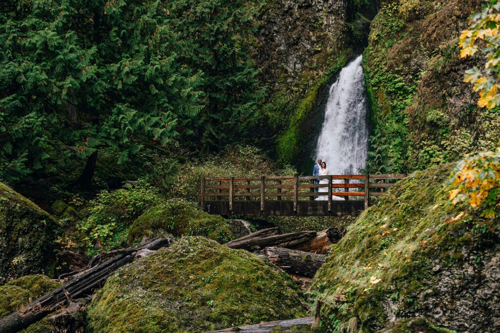 Beautiful couple standing on a bridge in their wedding clothes with a waterfall behind them in Oregon.