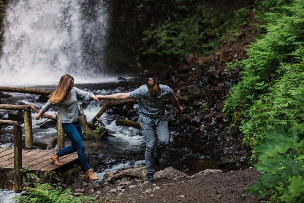 A man helps a woman off a bridge over a creek with a waterfall in the background.
