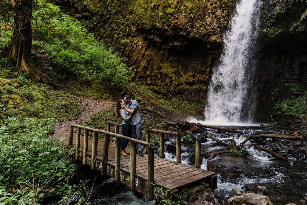 A couple stands on a bridge over a creek with a waterfall behind them.