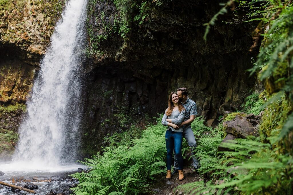 A man embraces a woman kissing her cheek standing next to lush green ferns with a waterfall cascading behind them.