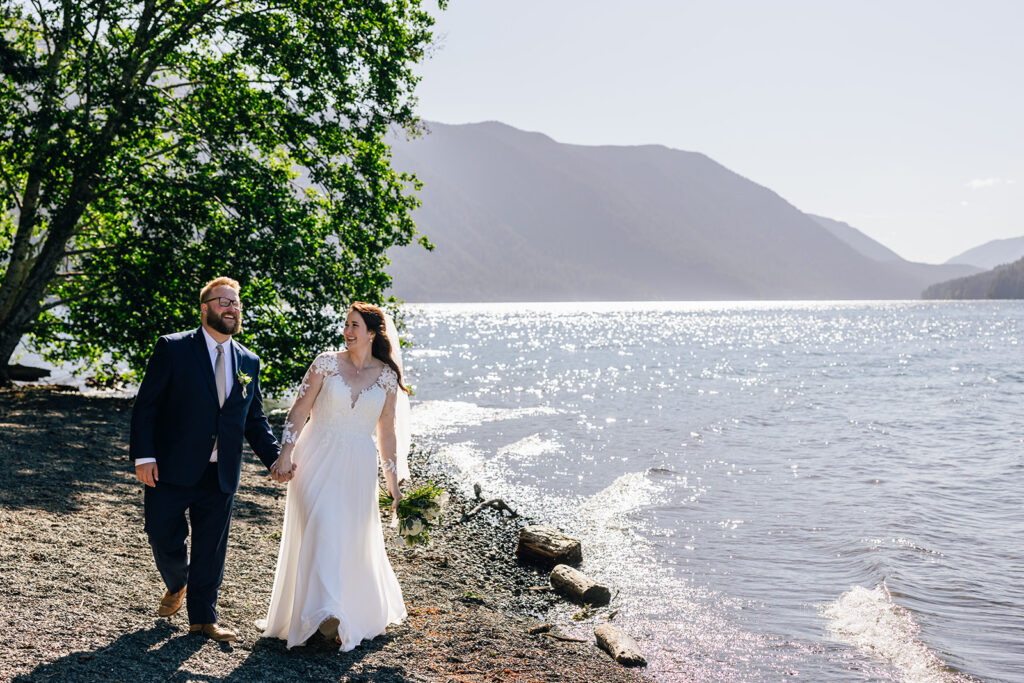 A wedding couple walks in the sun along the bank of Lake Crescent on their wedding day. 