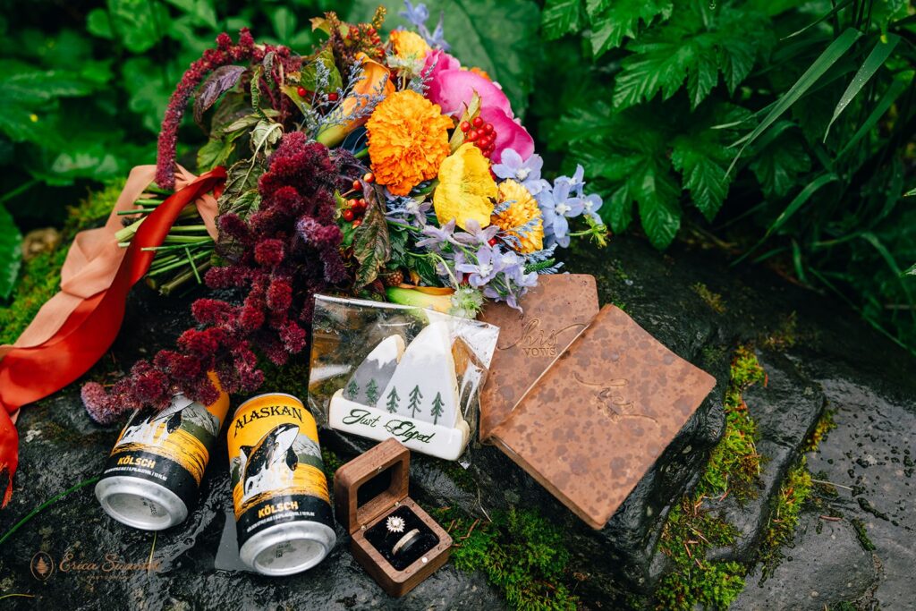 adventure elopement detail flatlay with the bouquet, vow books, beers, and rings